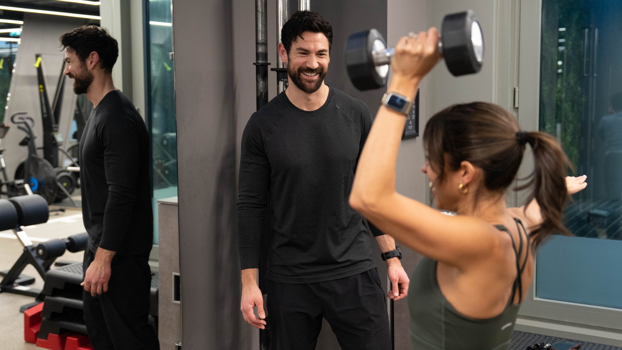 A woman pressing a dumbbell overhead while a trainer watches