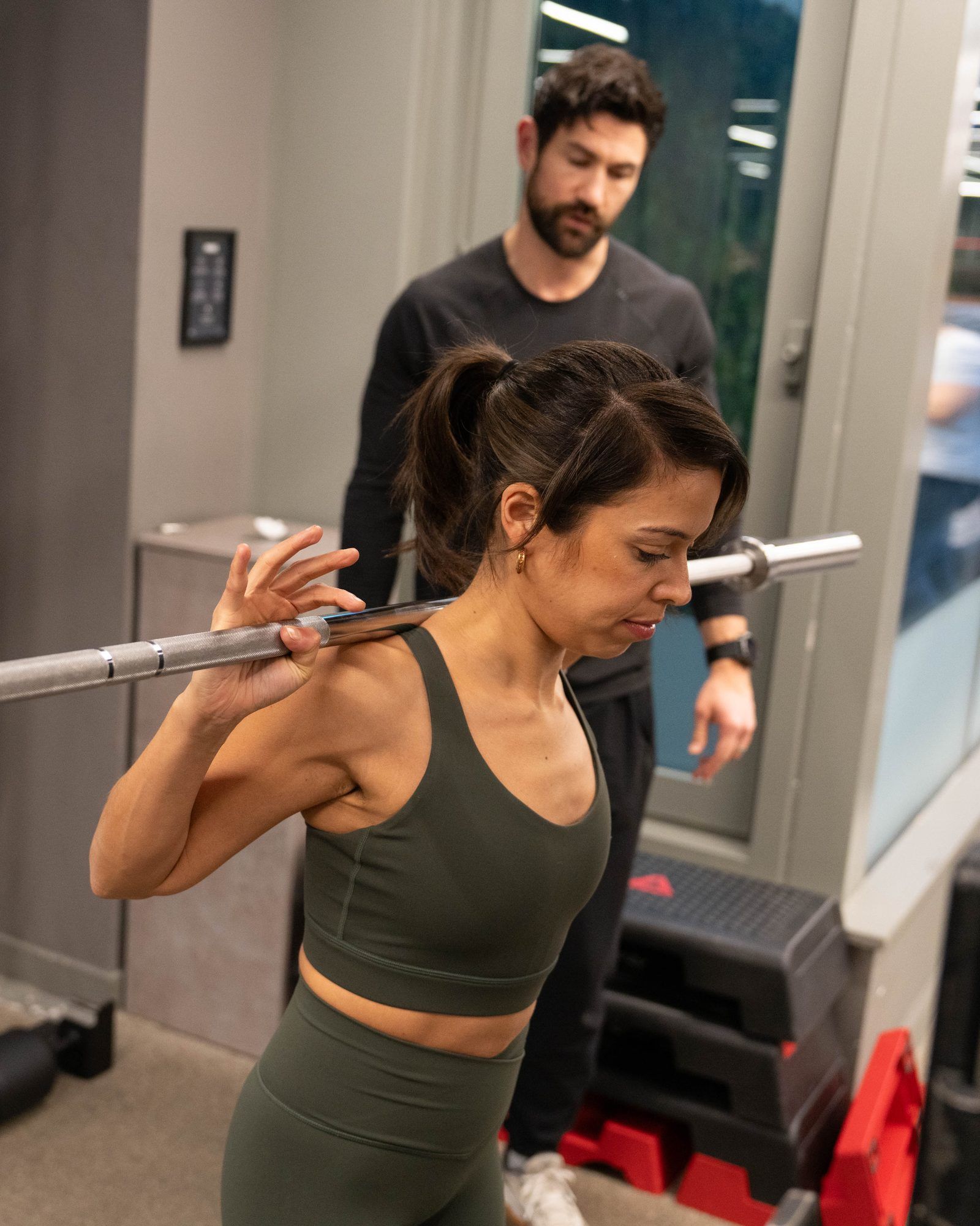 A woman setting a barbell across her shoulders with a coach standing behind her on the training floor