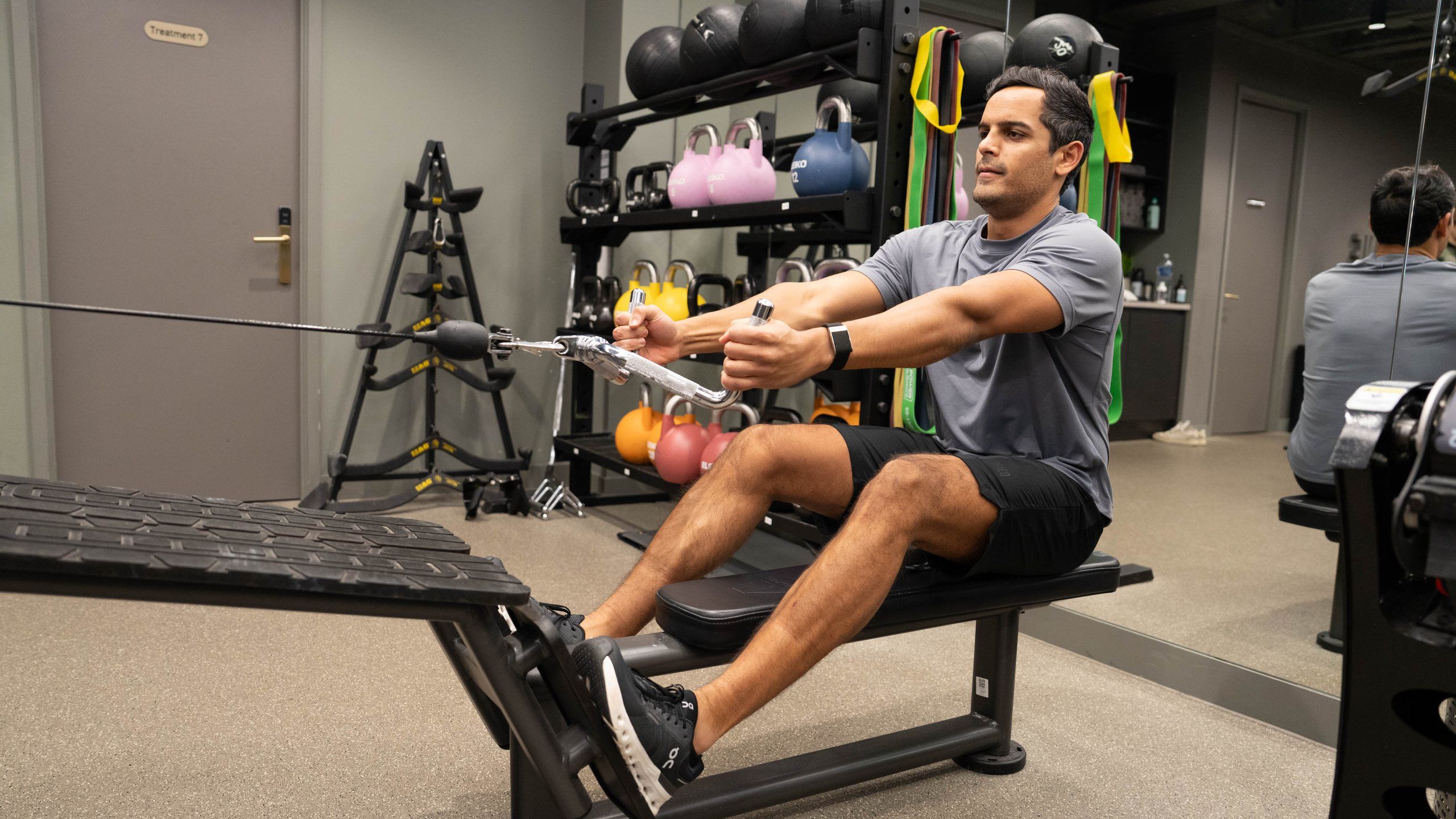 A man doing a seated cable row in front of a rack of kettlebells and resistance bands