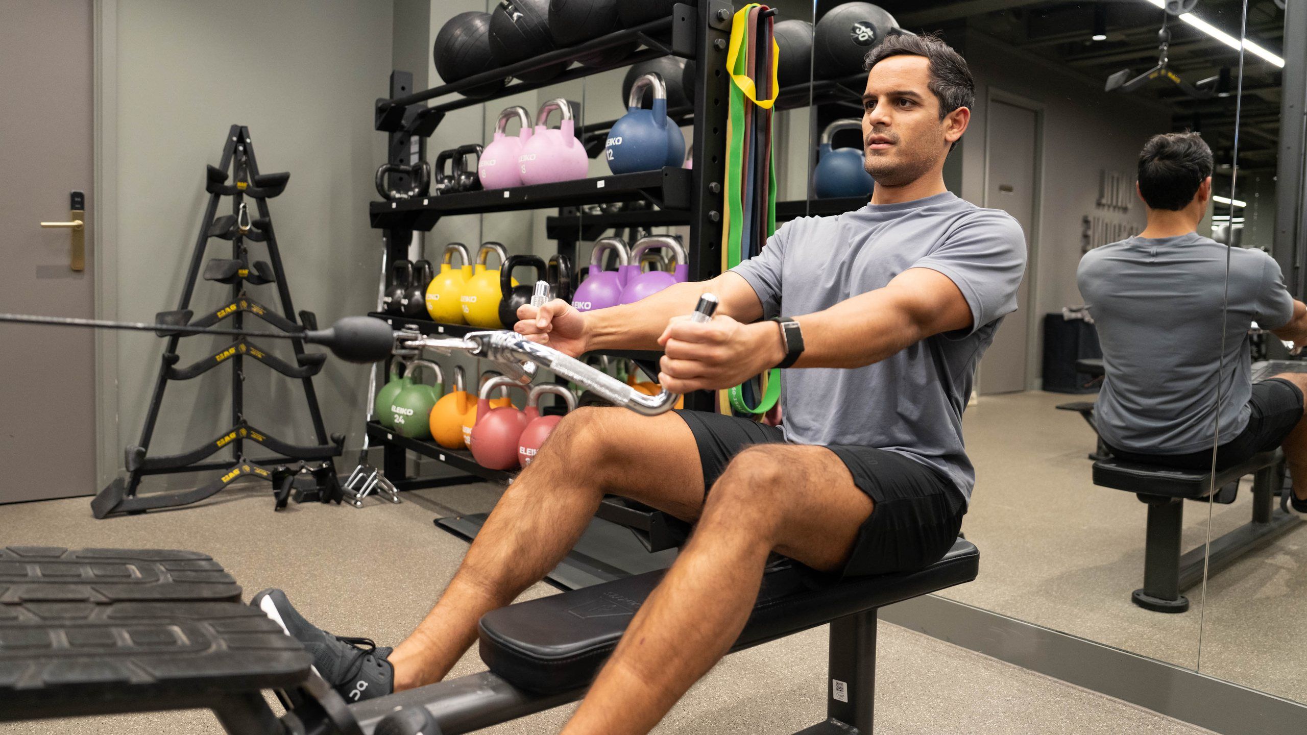 A man doing a seated cable row with kettlebells racked on the wall behind him