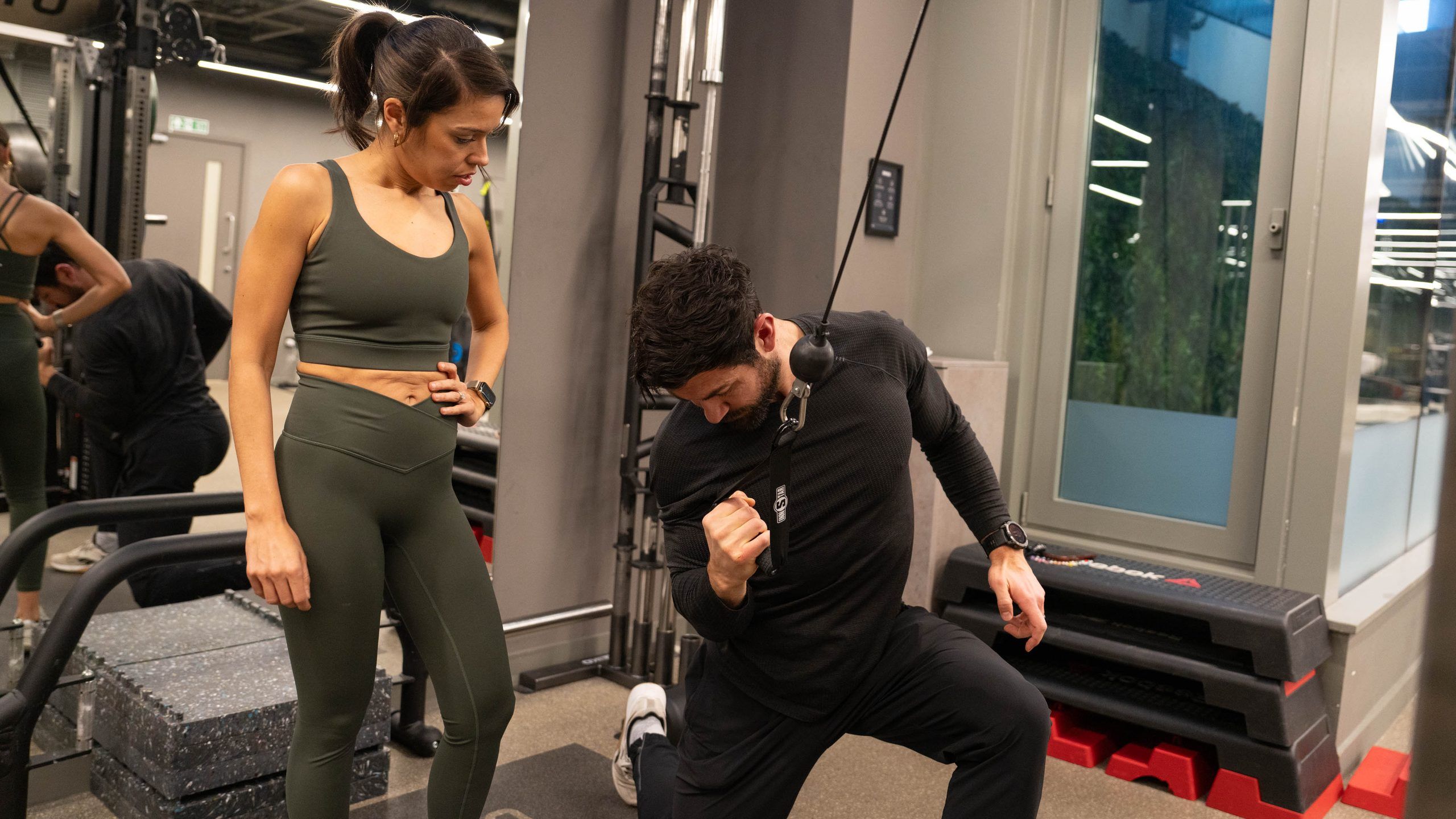 Client watching as a trainer demonstrates a kneeling cable exercise