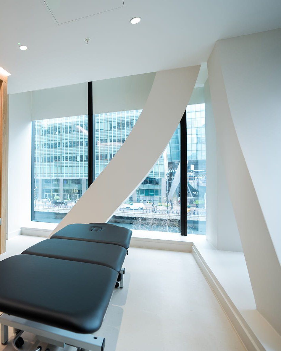 A treatment couch beside a tall window in a room at UNTIL Canary Wharf