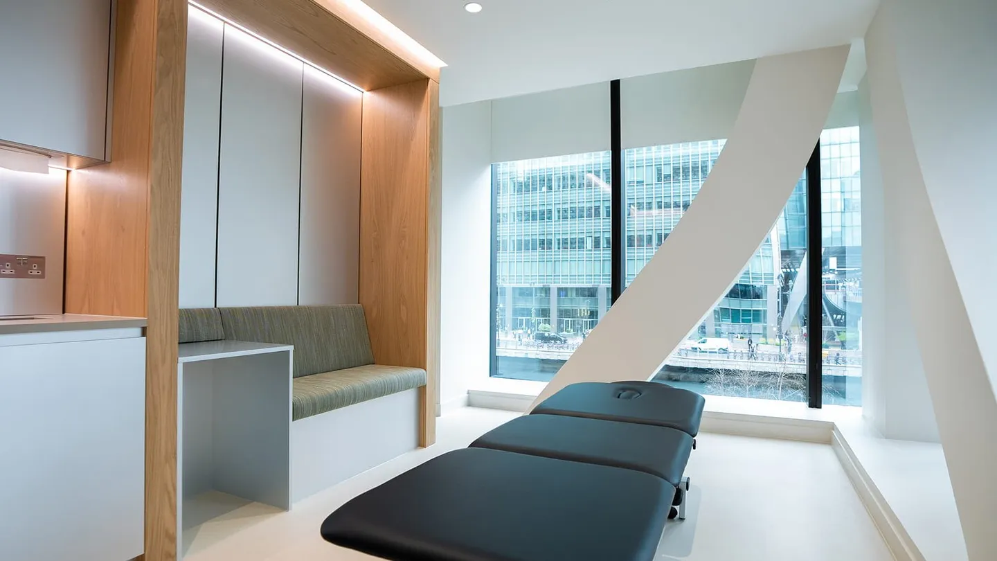 A treatment couch in a room at UNTIL Canary Wharf, with a tall window looking out on office towers