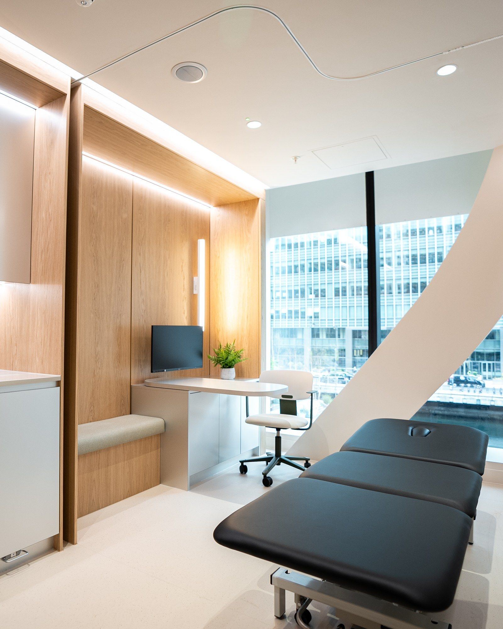 A physiotherapy treatment room at UNTIL Canary Wharf, with a treatment plinth, a consulting desk and a window over the dock.