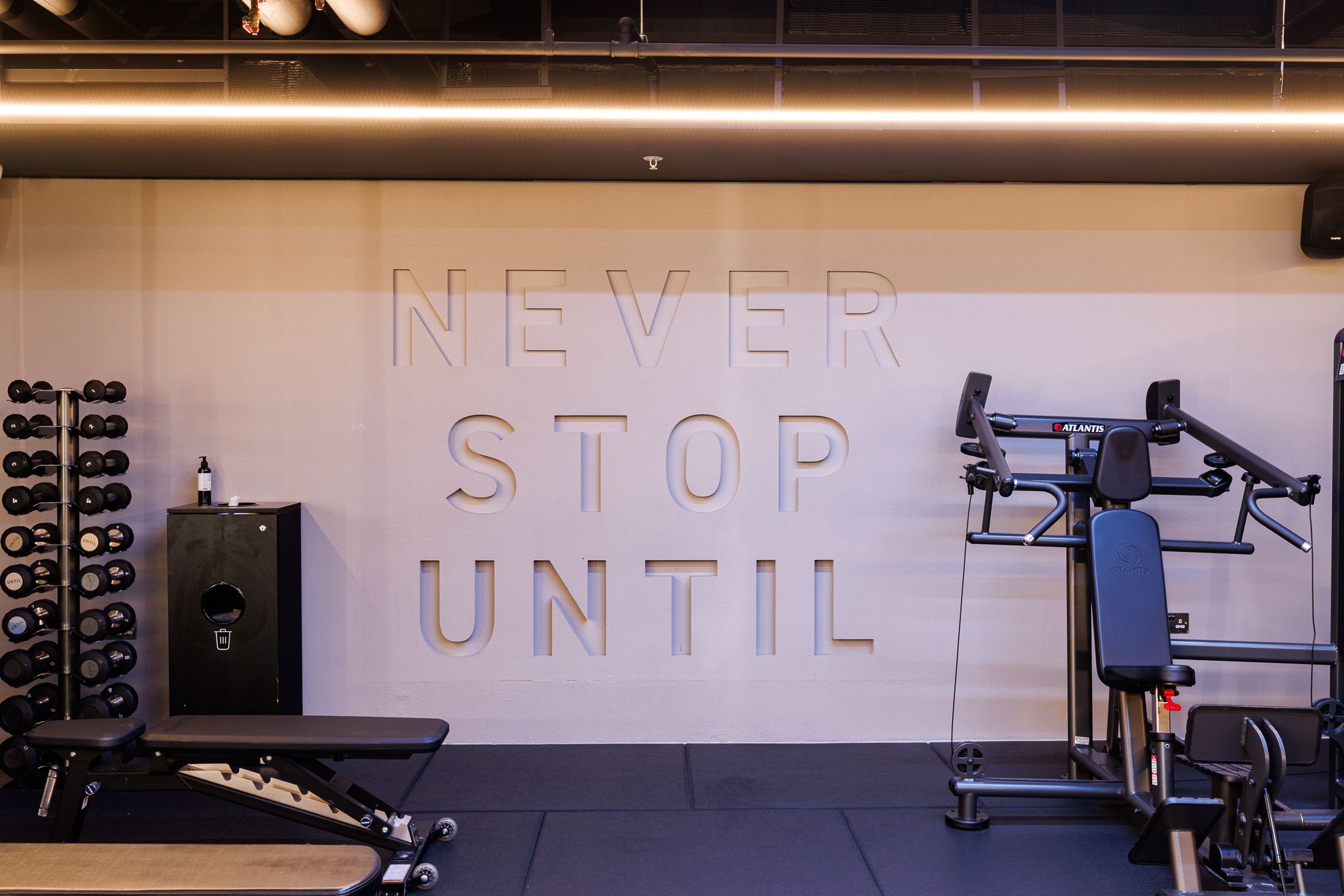 The "Never Stop Until" lettering on the wall at UNTIL Soho, beside cable machines and a bench