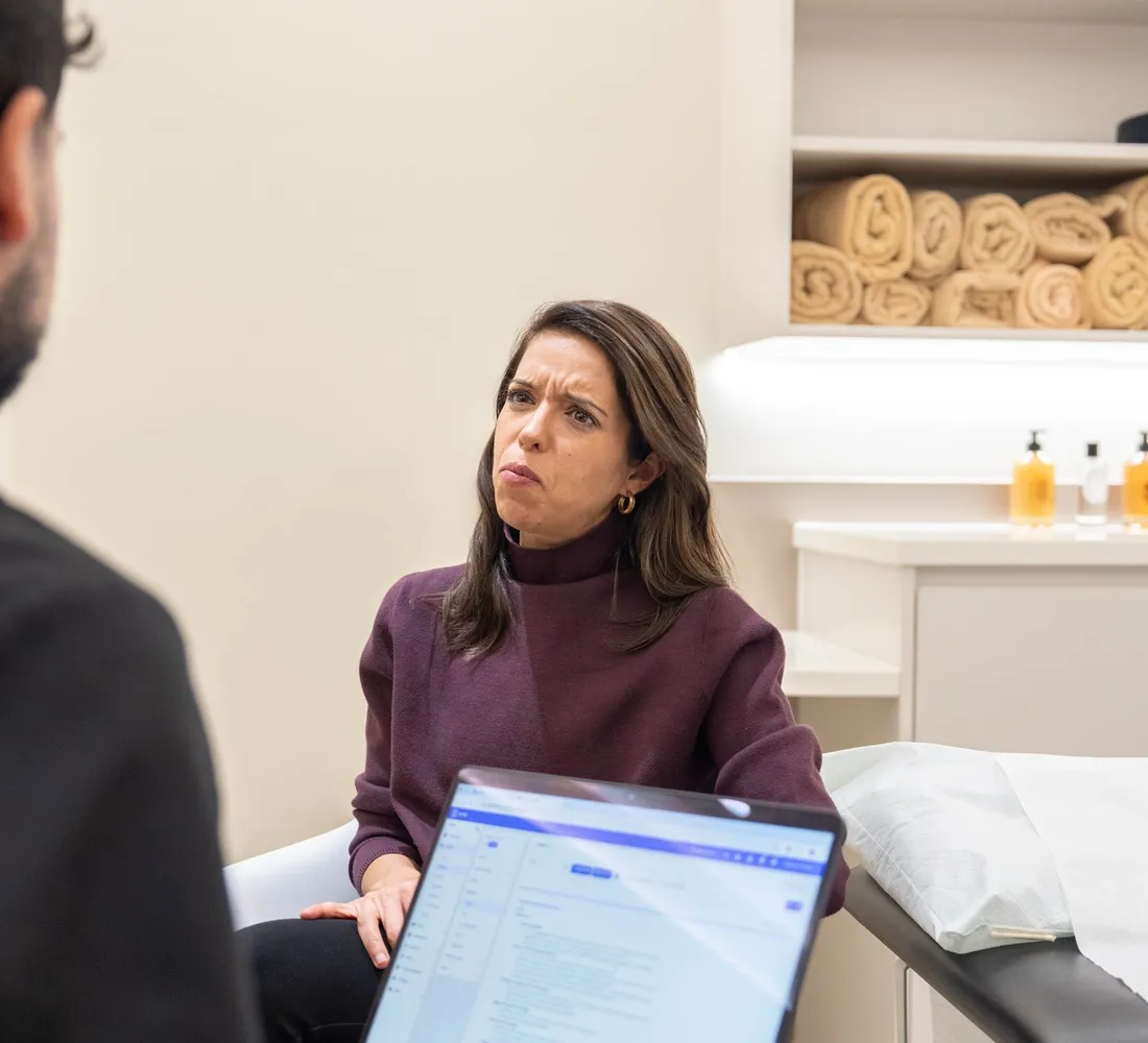 A client in a clinic room talks with a clinician who has notes open on a laptop