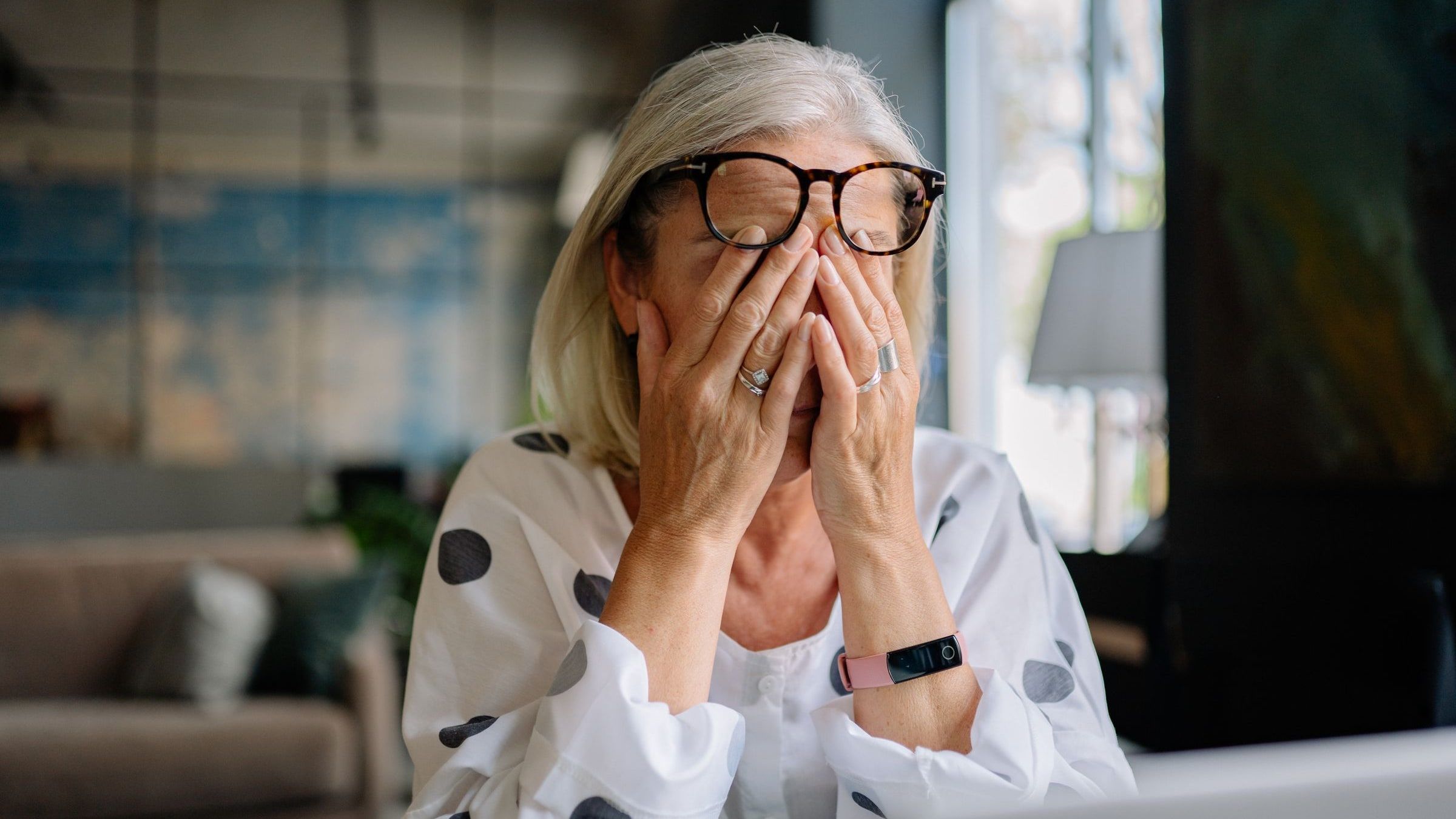 A woman in her forties sitting at a laptop at home, glasses pushed up as she rubs her closed eyes with both hands