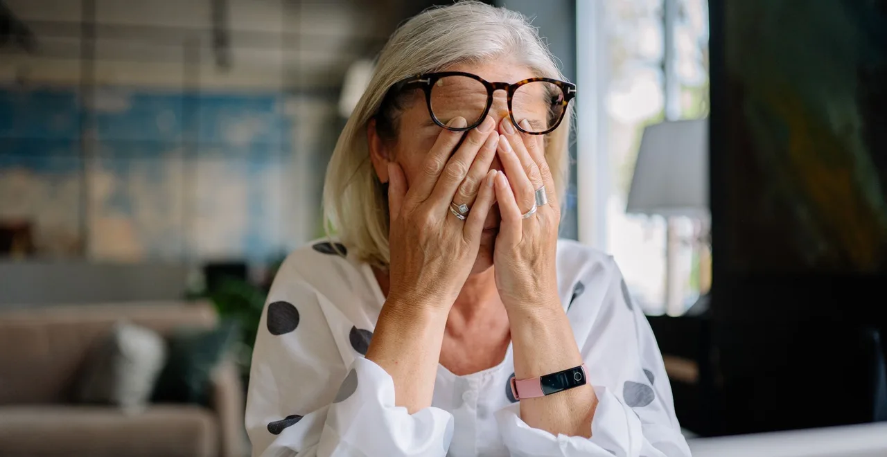 A woman in her forties sitting at a laptop at home, glasses pushed up as she rubs her closed eyes with both hands