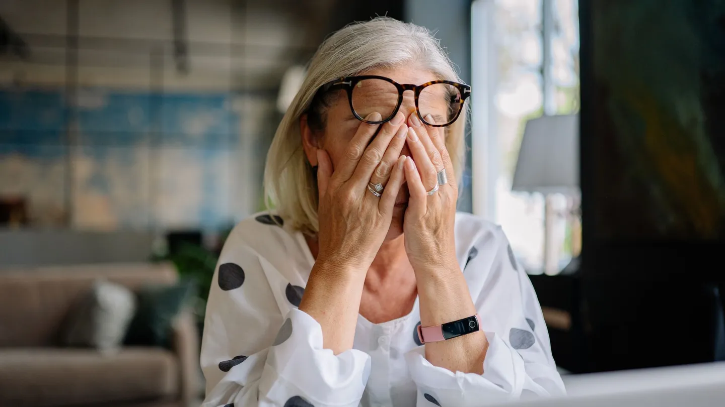 A woman in her forties sitting at a laptop at home, glasses pushed up as she rubs her closed eyes with both hands