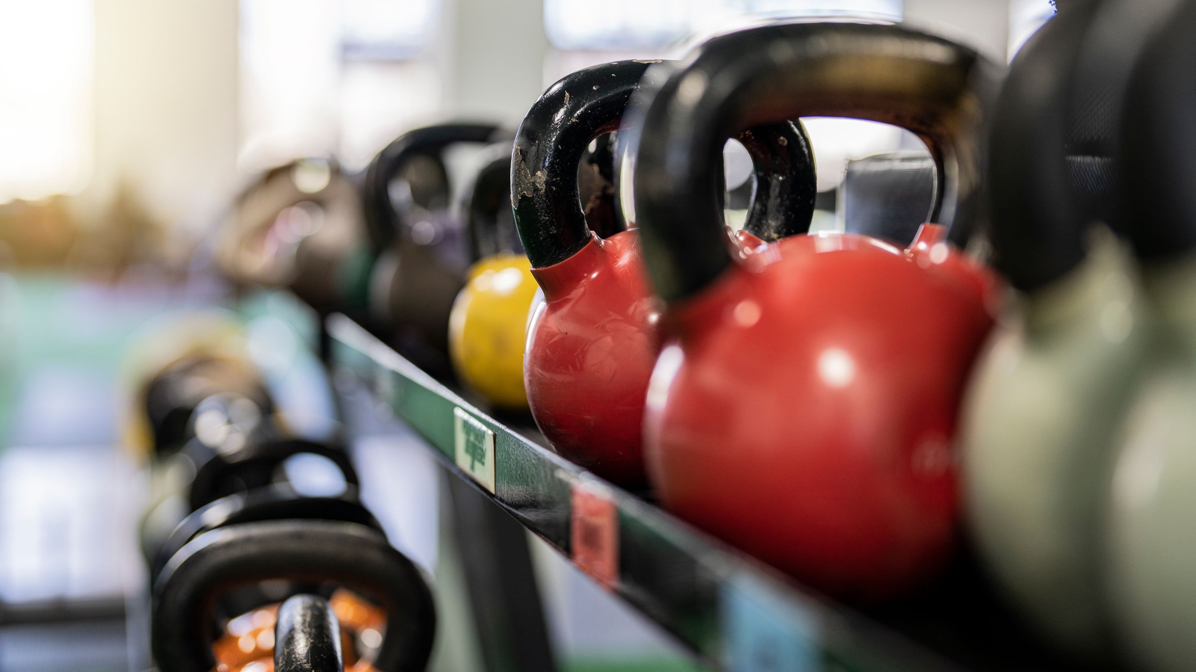 A row of kettlebells on a gym rack, two red bells in focus in the foreground with dumbbells racked behind them