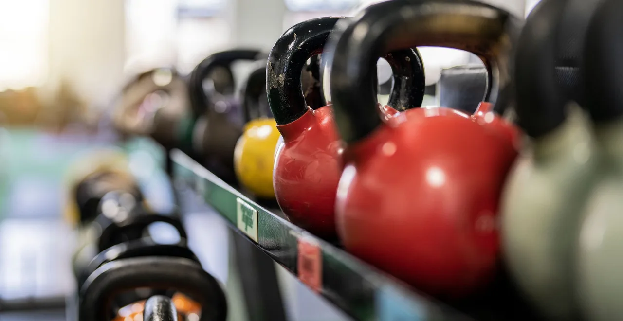 A row of kettlebells on a gym rack, two red bells in focus in the foreground with dumbbells racked behind them