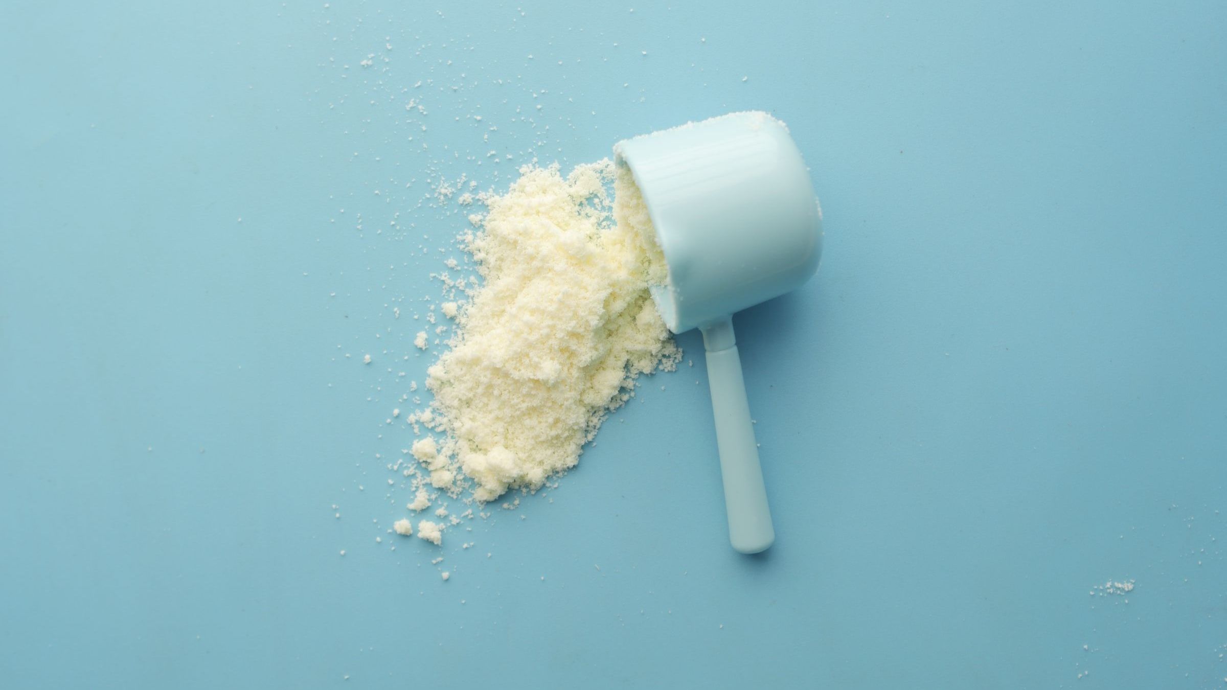A white plastic supplement scoop tipped on its side on a pale blue background, with creatine powder spilling out of it