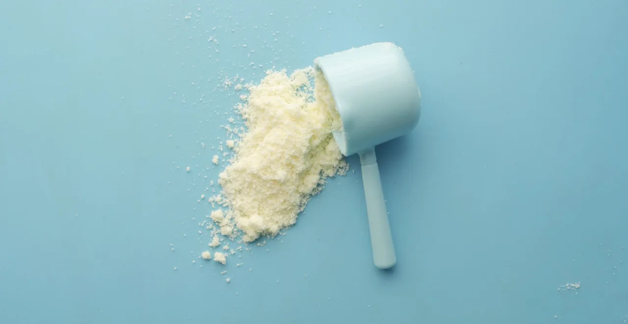 A white plastic supplement scoop tipped on its side on a pale blue background, with creatine powder spilling out of it