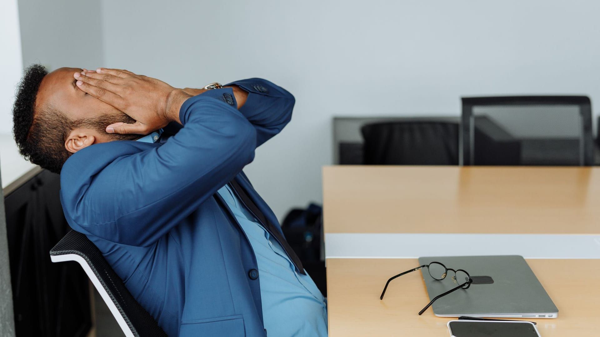 Man in a suit leaning back at his office desk with his hands behind his head, stretching out his neck and shoulders after desk work