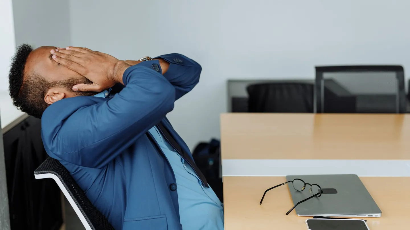 Man in a suit leaning back at his office desk with his hands behind his head, stretching out his neck and shoulders after desk work