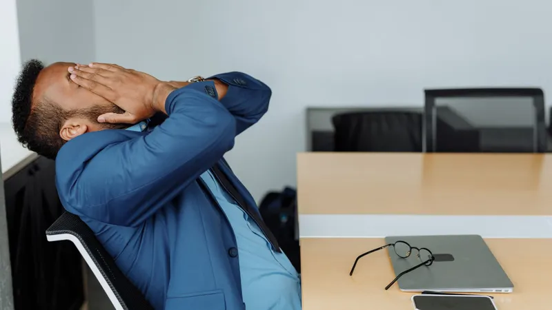 Man in a suit leaning back at his office desk with his hands behind his head, stretching out his neck and shoulders after desk work