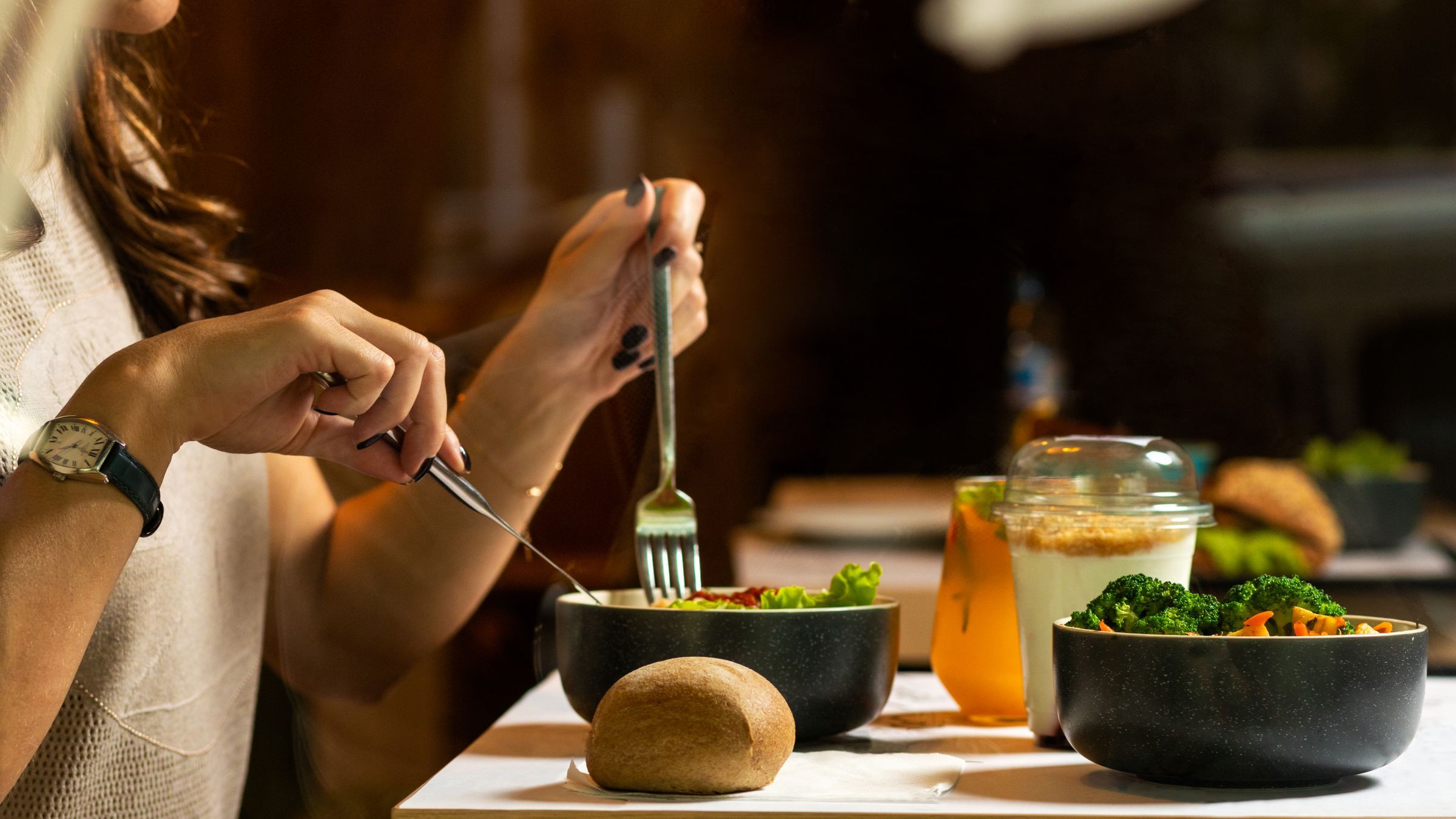 A woman wearing a wristwatch eats a meal with a knife and fork, with bowls of salad and broccoli, a yoghurt pot and a bread roll on the table