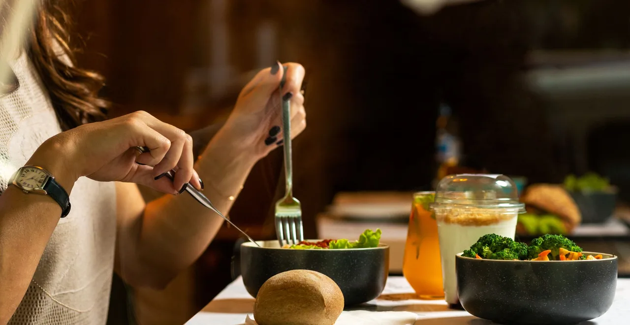 A woman wearing a wristwatch eats a meal with a knife and fork, with bowls of salad and broccoli, a yoghurt pot and a bread roll on the table