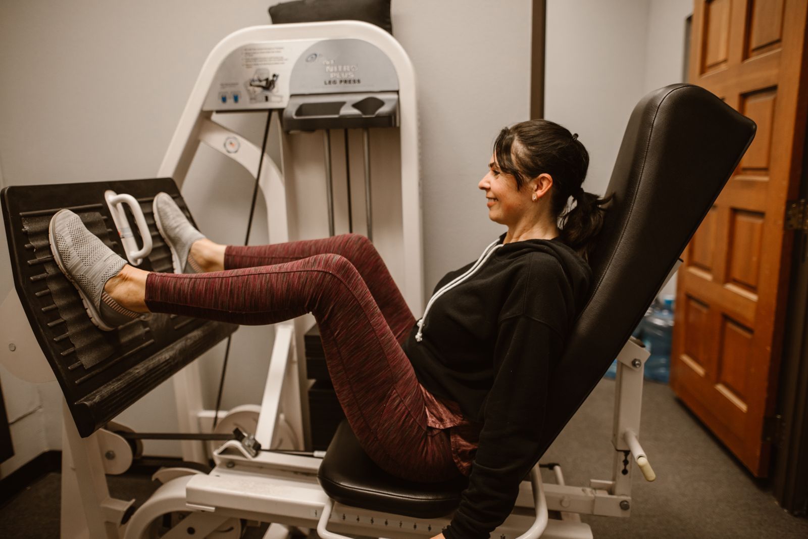 A woman in her forties pressing a weighted leg press machine in a gym, seated back against the pad with her legs extended