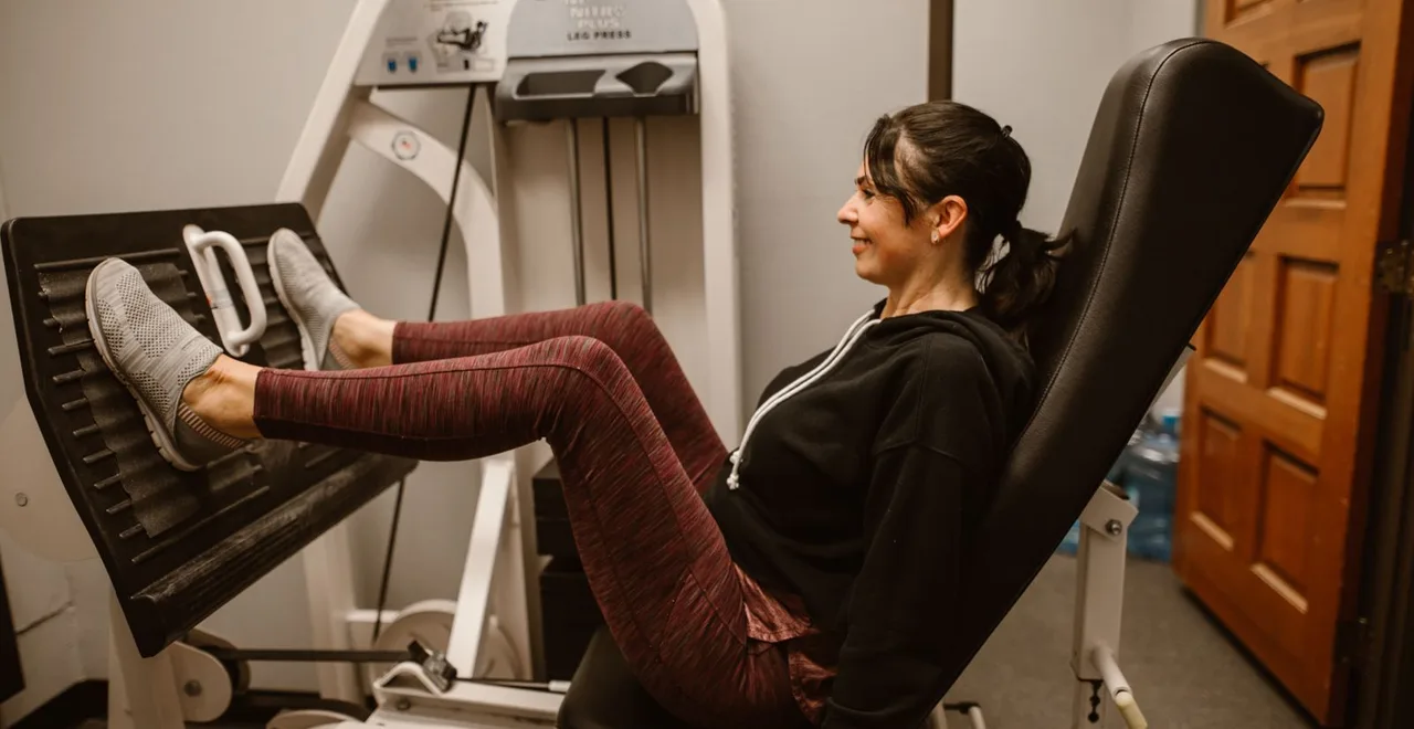 A woman in her forties pressing a weighted leg press machine in a gym, seated back against the pad with her legs extended