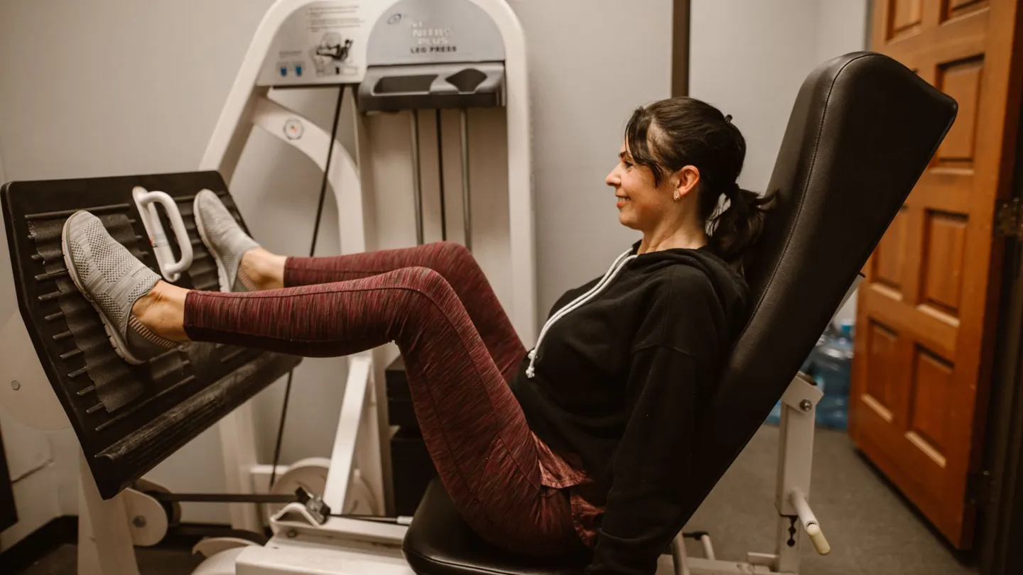 A woman in her forties pressing a weighted leg press machine in a gym, seated back against the pad with her legs extended