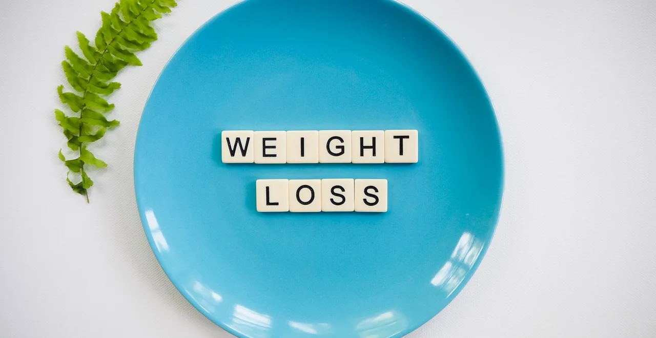 Letter tiles spelling WEIGHT LOSS arranged on a turquoise plate, photographed from above on a white surface beside a green fern frond