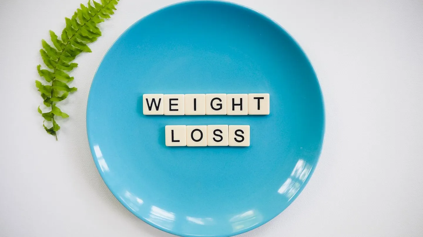 Letter tiles spelling WEIGHT LOSS arranged on a turquoise plate, photographed from above on a white surface beside a green fern frond