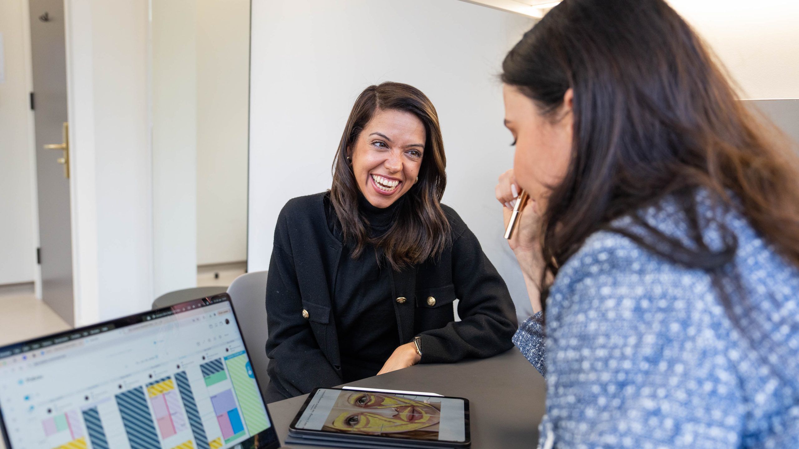 A smiling client talks with a practitioner across a desk, with a skin analysis open on a laptop and a tablet