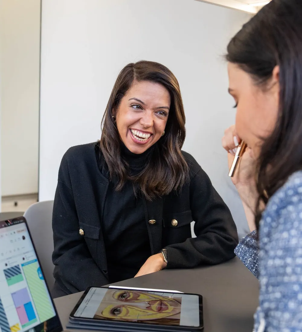 A smiling client talks with a practitioner across a desk, with a skin analysis open on a laptop and a tablet