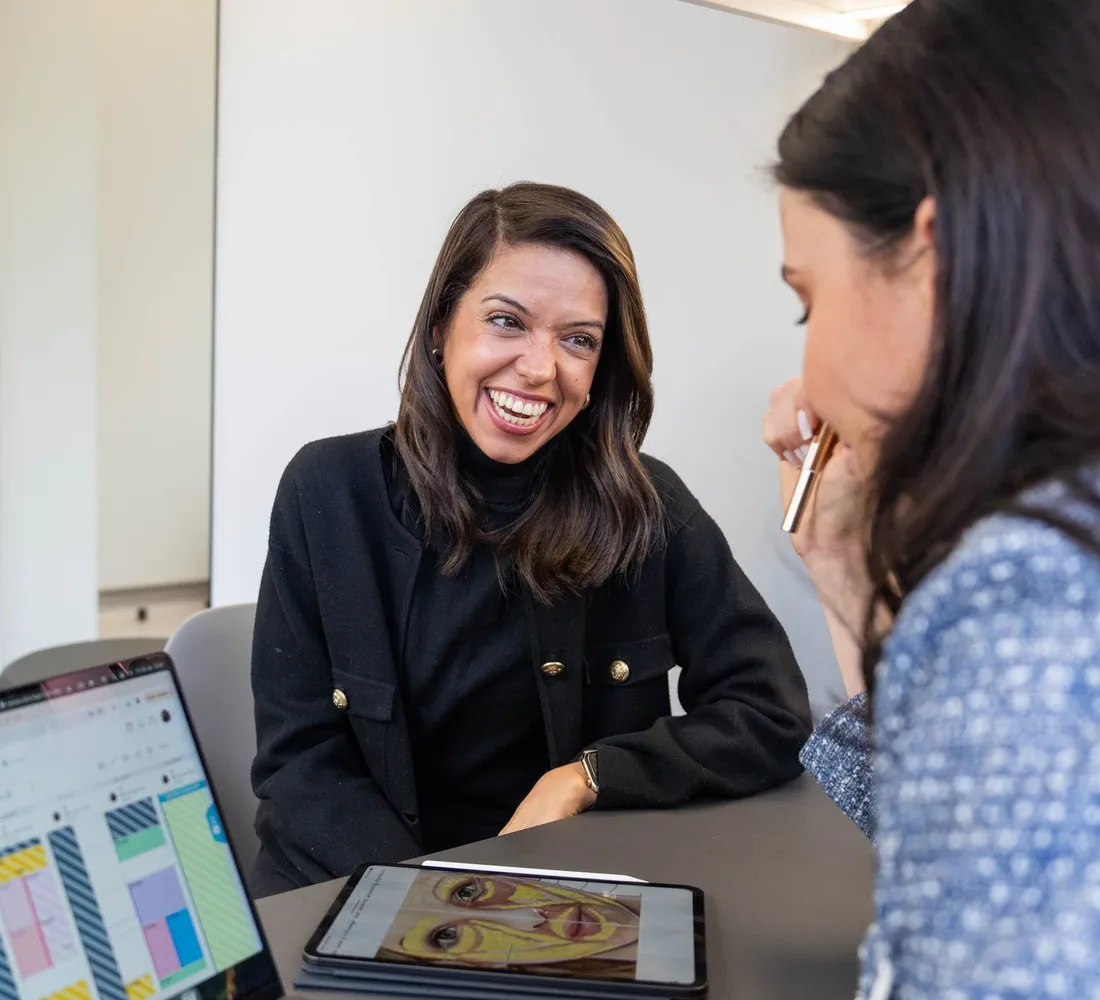 A smiling client talks with a practitioner across a desk, with a skin analysis open on a laptop and a tablet