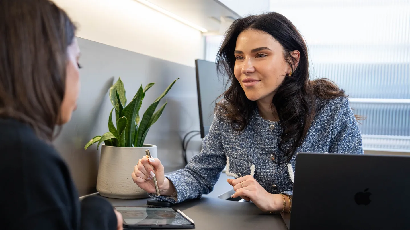 A practitioner holding a stylus smiles as she talks with a client across a desk, with a laptop and a tablet beside her