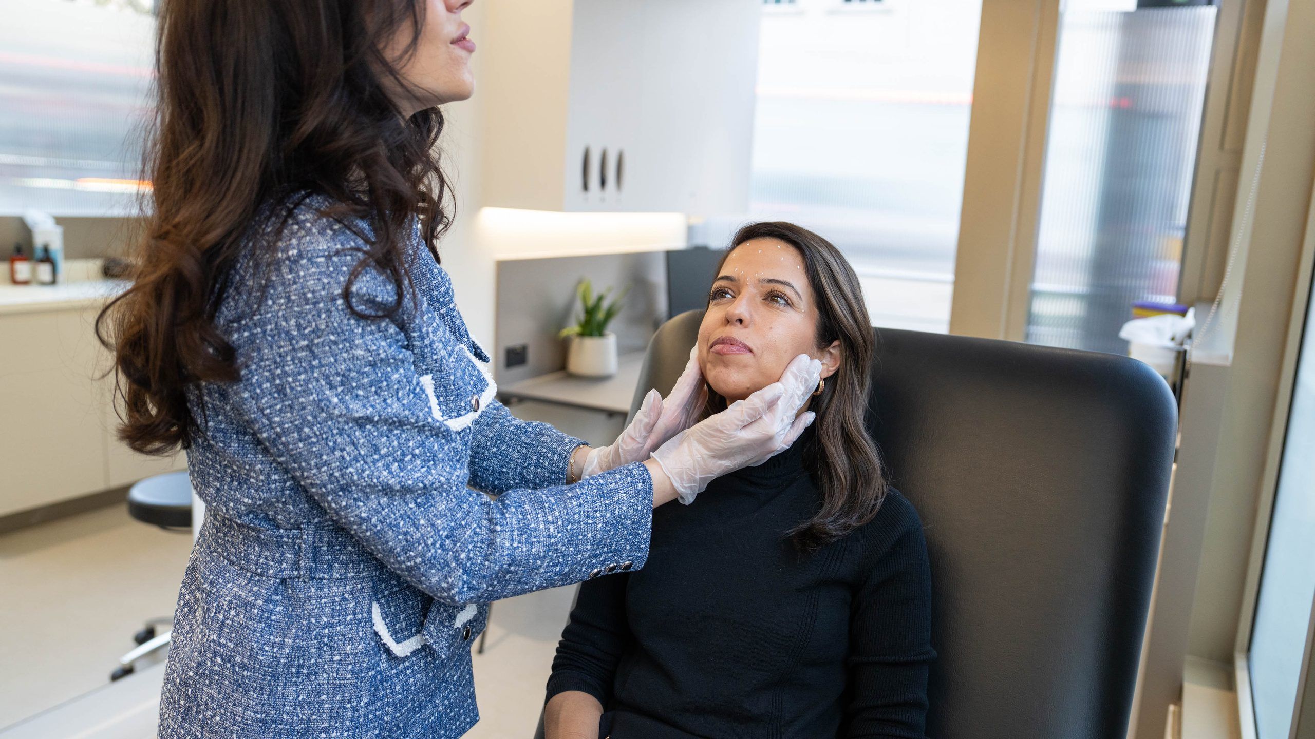 A gloved practitioner holds a client’s face to look at her cheeks and jawline in daylight