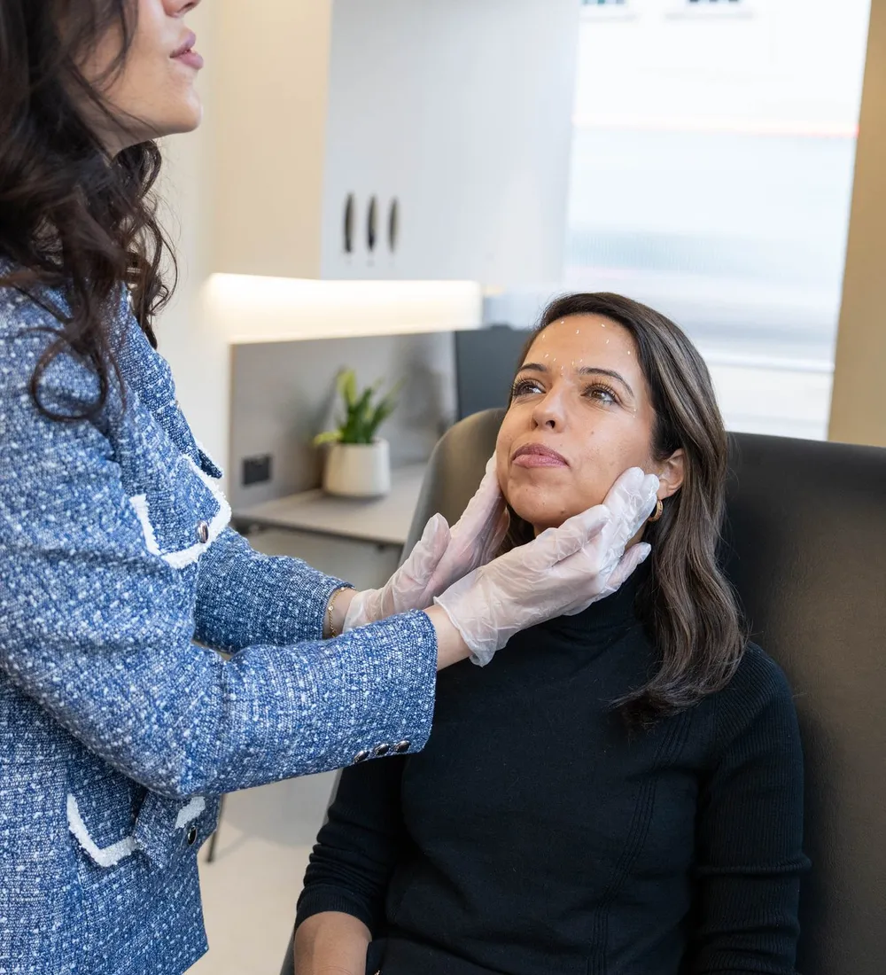 A gloved practitioner holds a client’s face to look at her cheeks and jawline in daylight