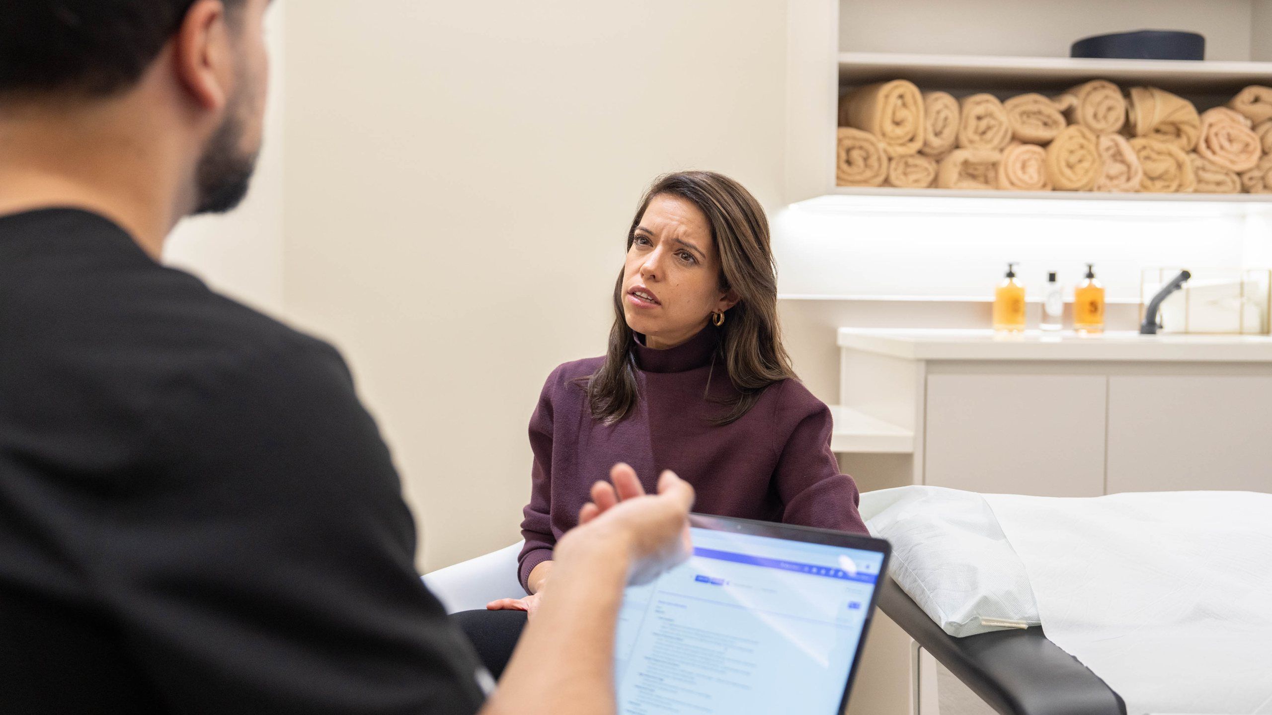 A client in a clinic room talks with a clinician, with notes open on a laptop