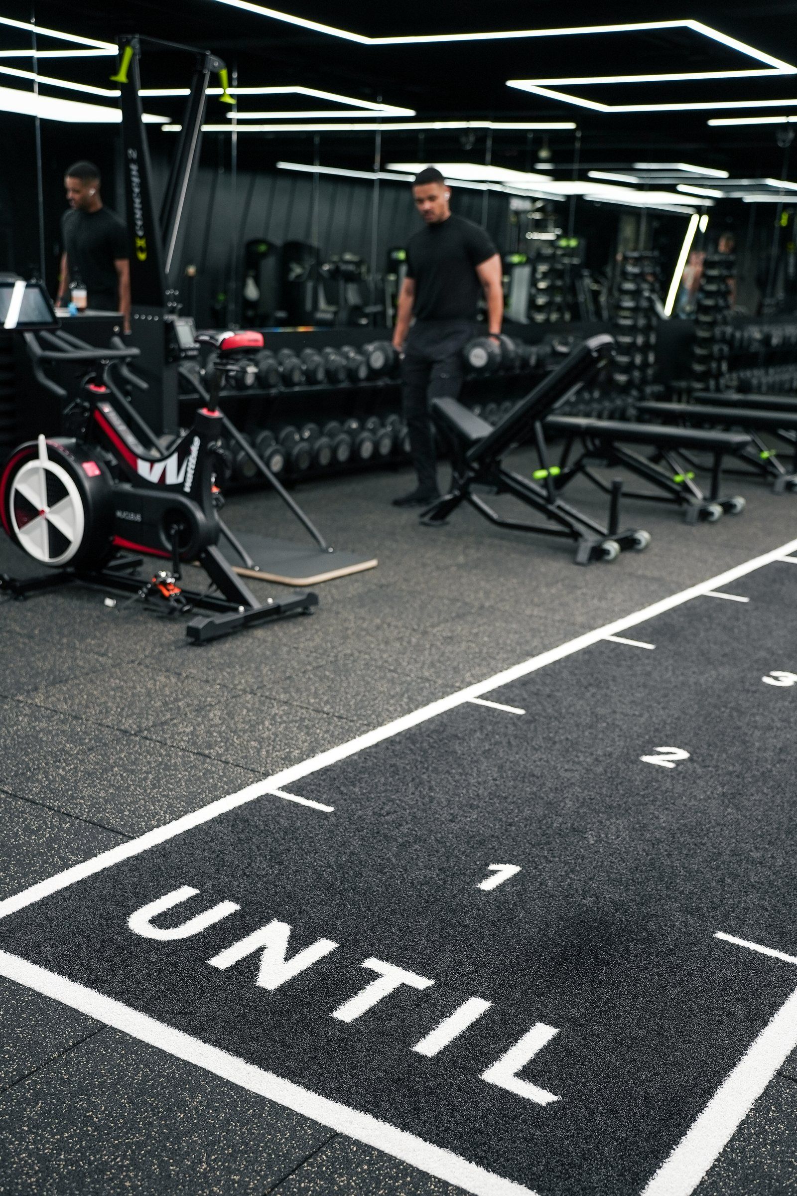The UNTIL gym floor with its lettering and marked lane, machines and benches behind
