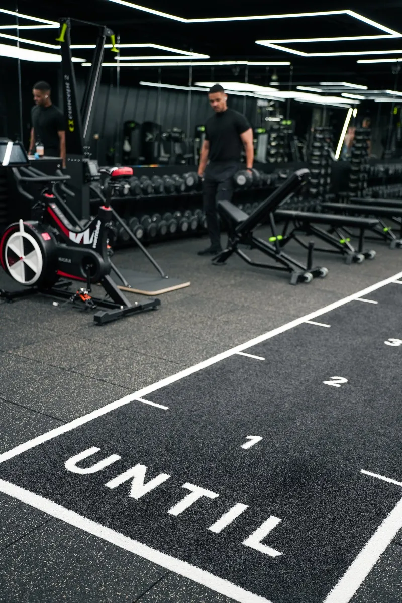 The UNTIL gym floor with its lettering and marked lane, machines and benches behind