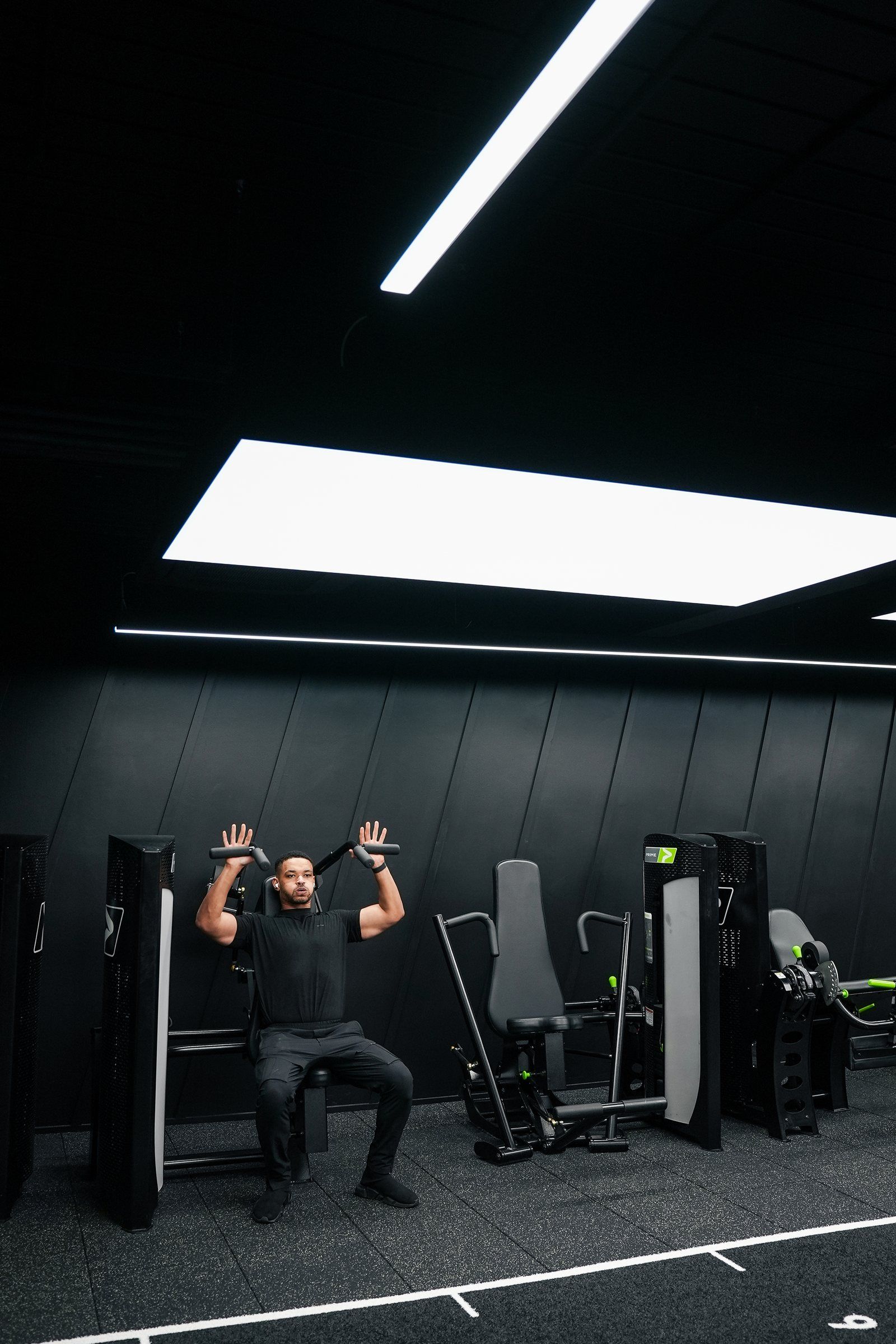 Steven Bartlett pressing overhead on a shoulder press machine, shot wide under the ceiling light panel