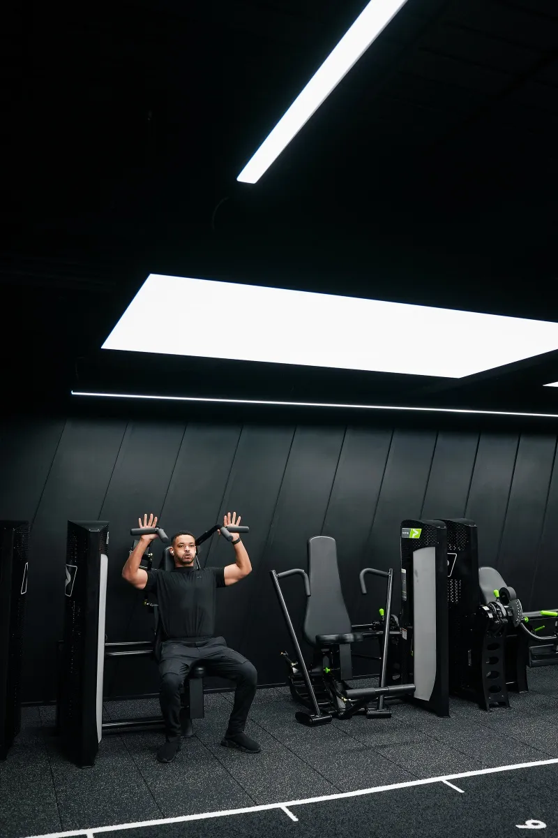Steven Bartlett pressing overhead on a shoulder press machine, shot wide under the ceiling light panel