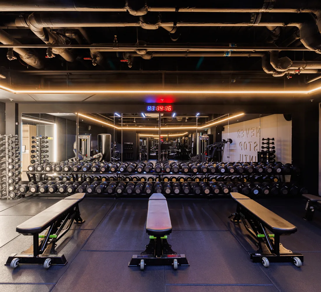 The dumbbell floor at UNTIL Soho, with a long dumbbell rack, adjustable benches and a timing clock above the mirrors