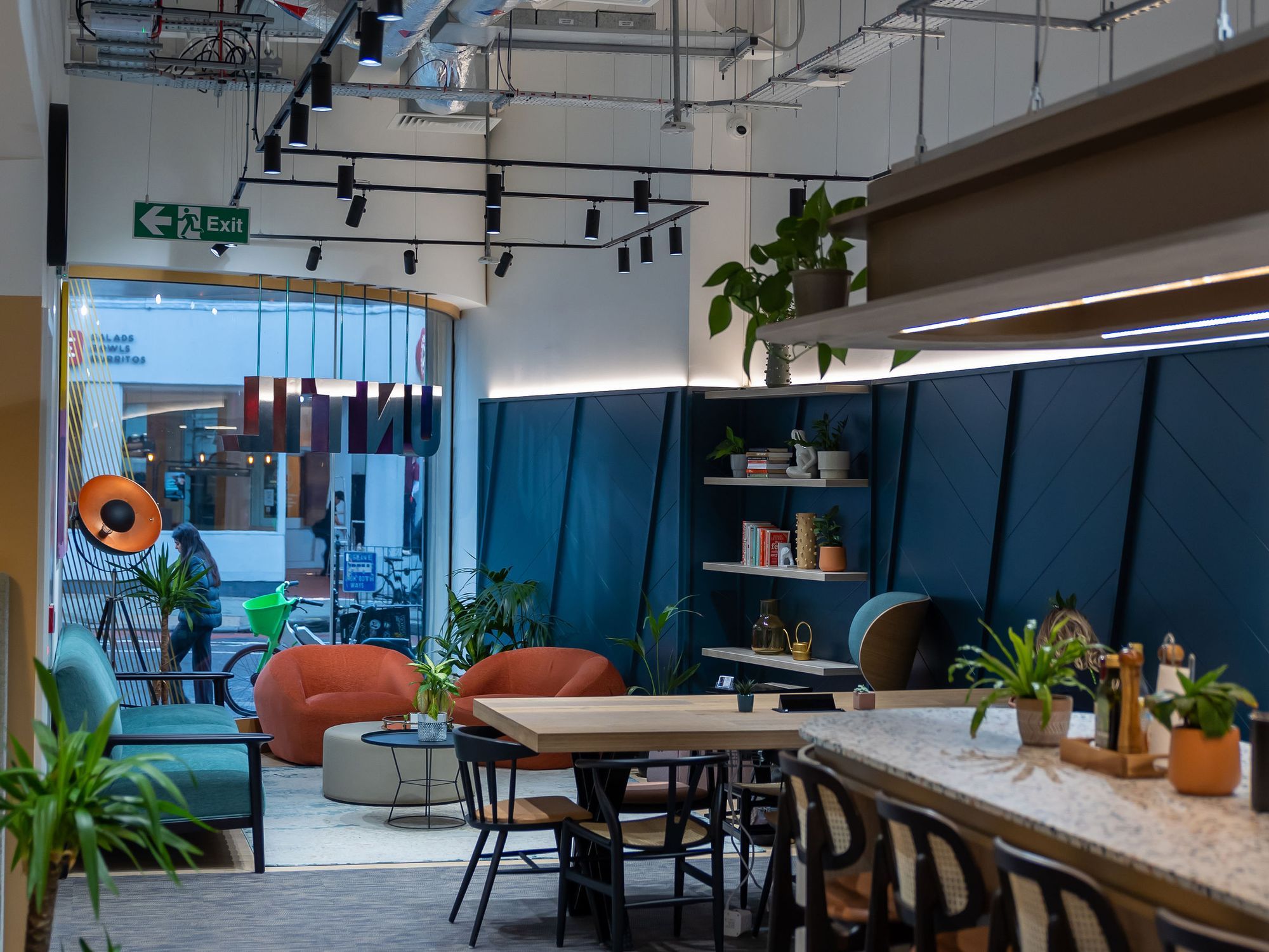 The social space at UNTIL Soho, with seating, plants and the street visible through the front windows.