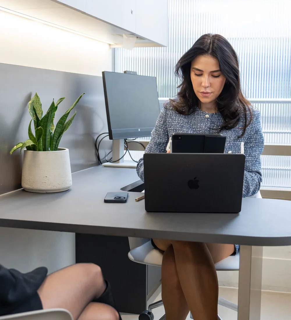 A practitioner works at a laptop by a window while a client sits across the desk listening
