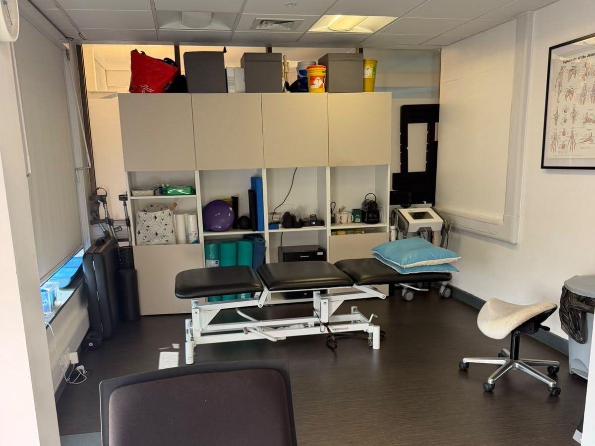 Treatment room at the Crouch End clinic: a height-adjustable plinth, rehab equipment on the shelving and an anatomy chart on the wall.