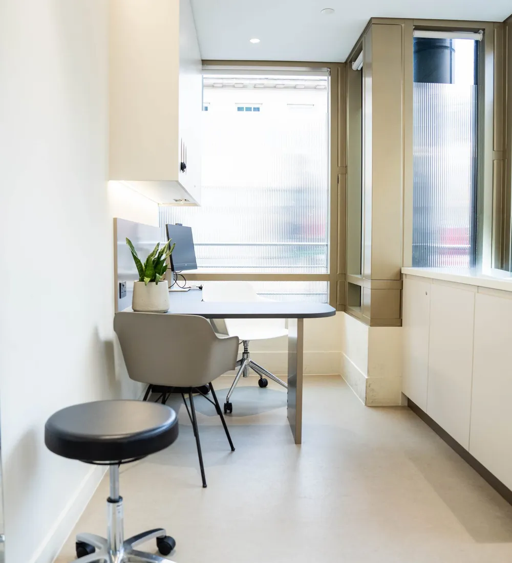 An empty treatment room with a stool, a chair by the window and pale wooden cabinets