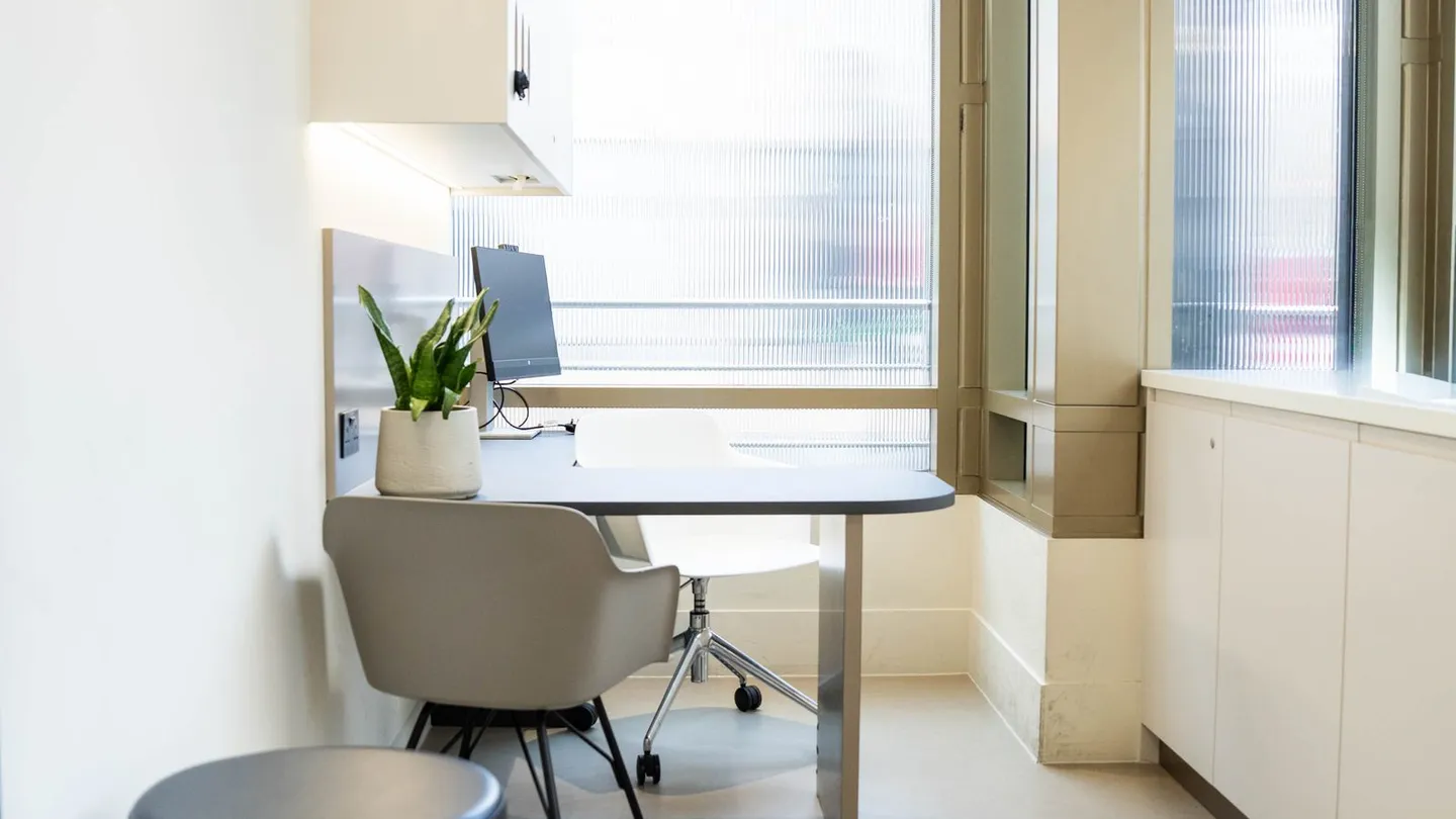An empty treatment room with a stool, a chair by the window and pale wooden cabinets