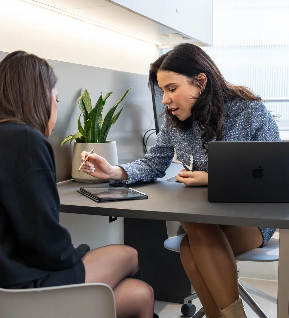 A practitioner points to a tablet on the desk while a client sits beside her by a large window